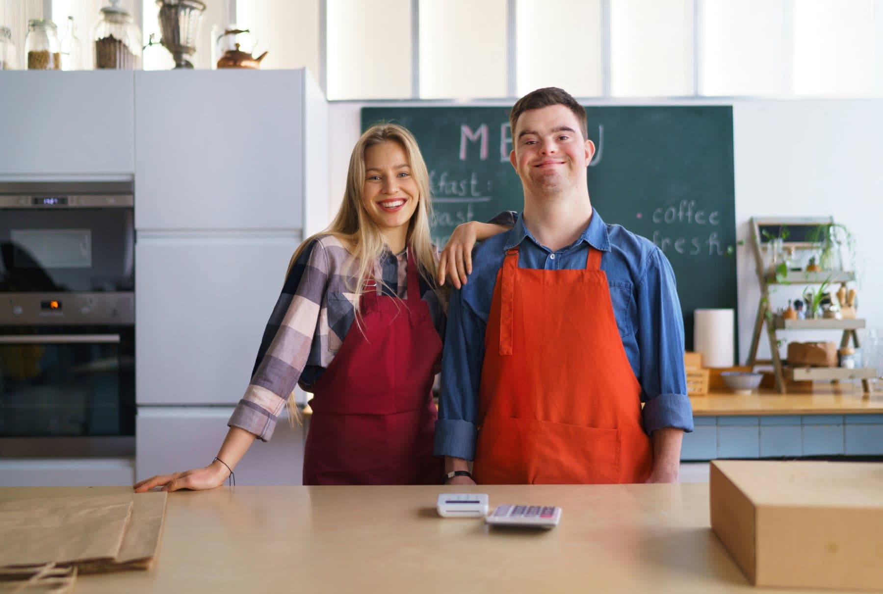 Two staff members in aprons smiling behind the counter of a café